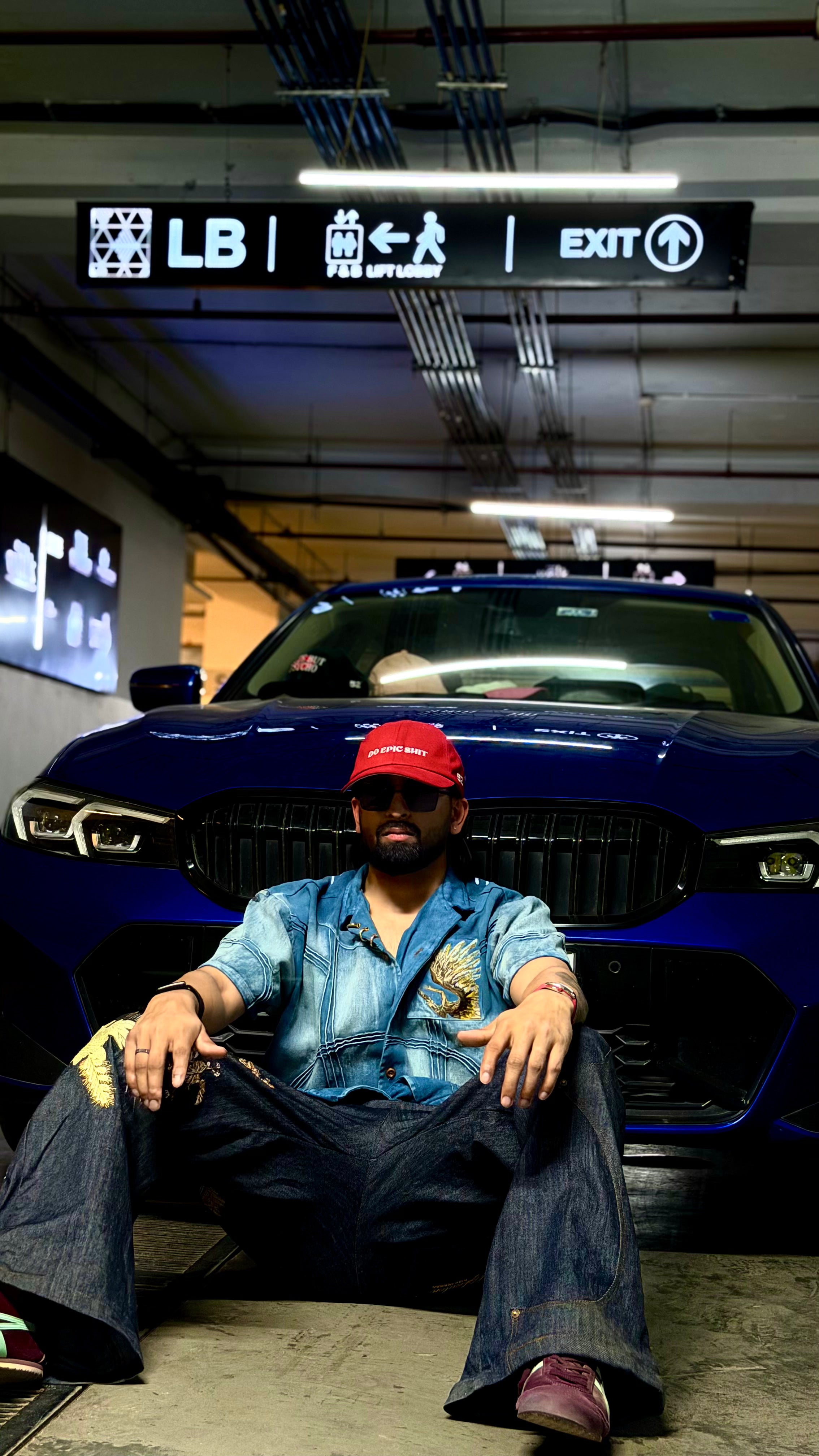 Person sitting in front of a blue car in a garage with an exit sign above.