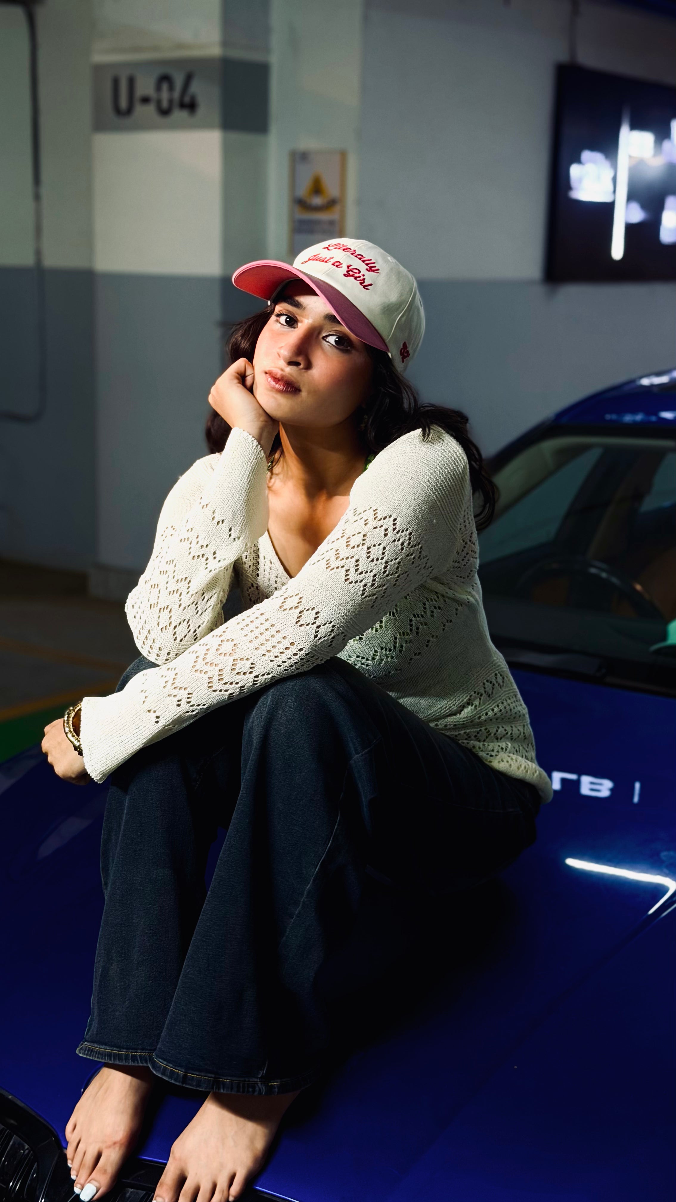 Woman sitting on a blue car wearing a white sweater and red cap inside a building.