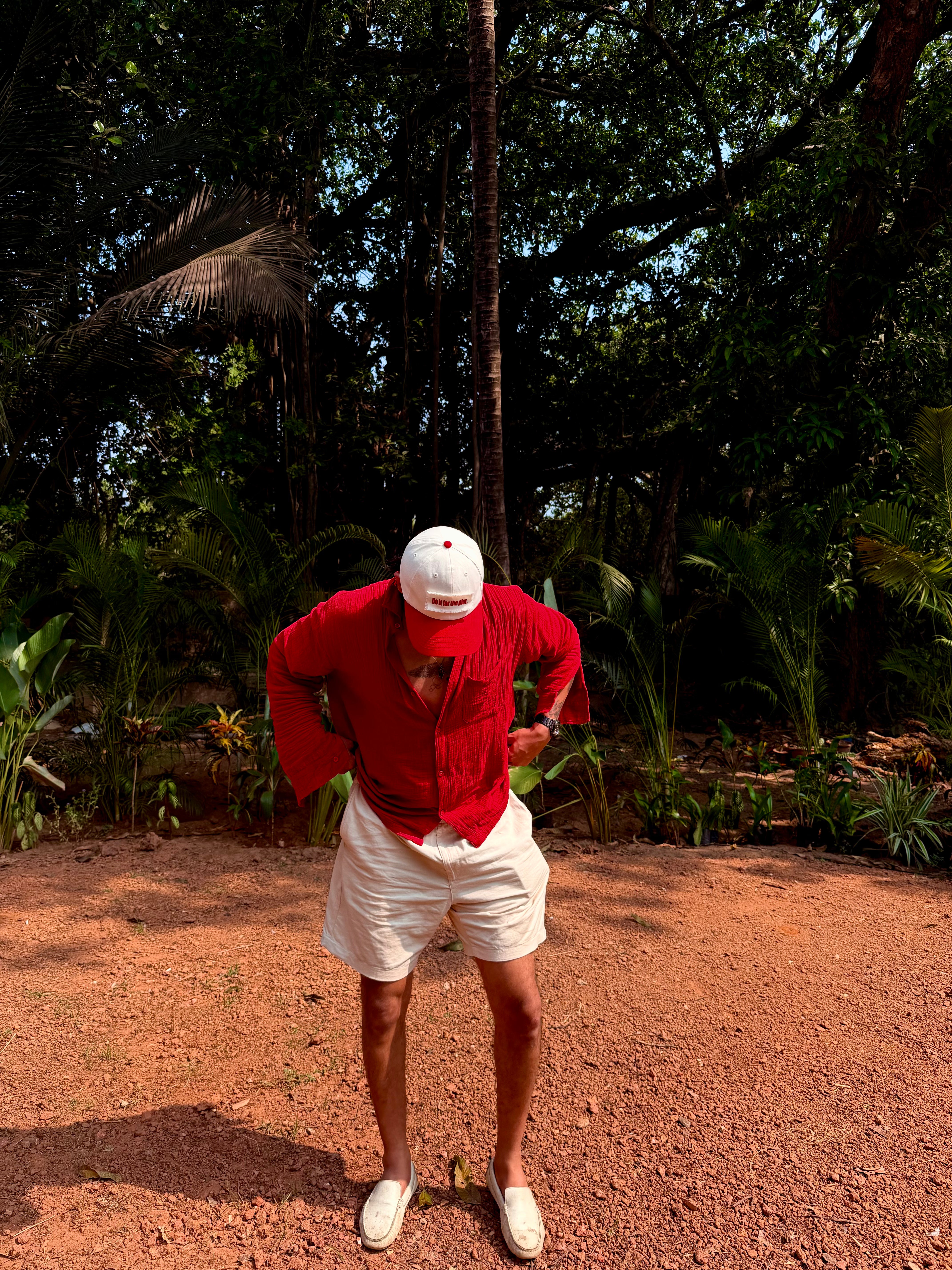 Person in red shirt and white shorts standing on a dirt path with trees in the background wearing do it for the plot cap