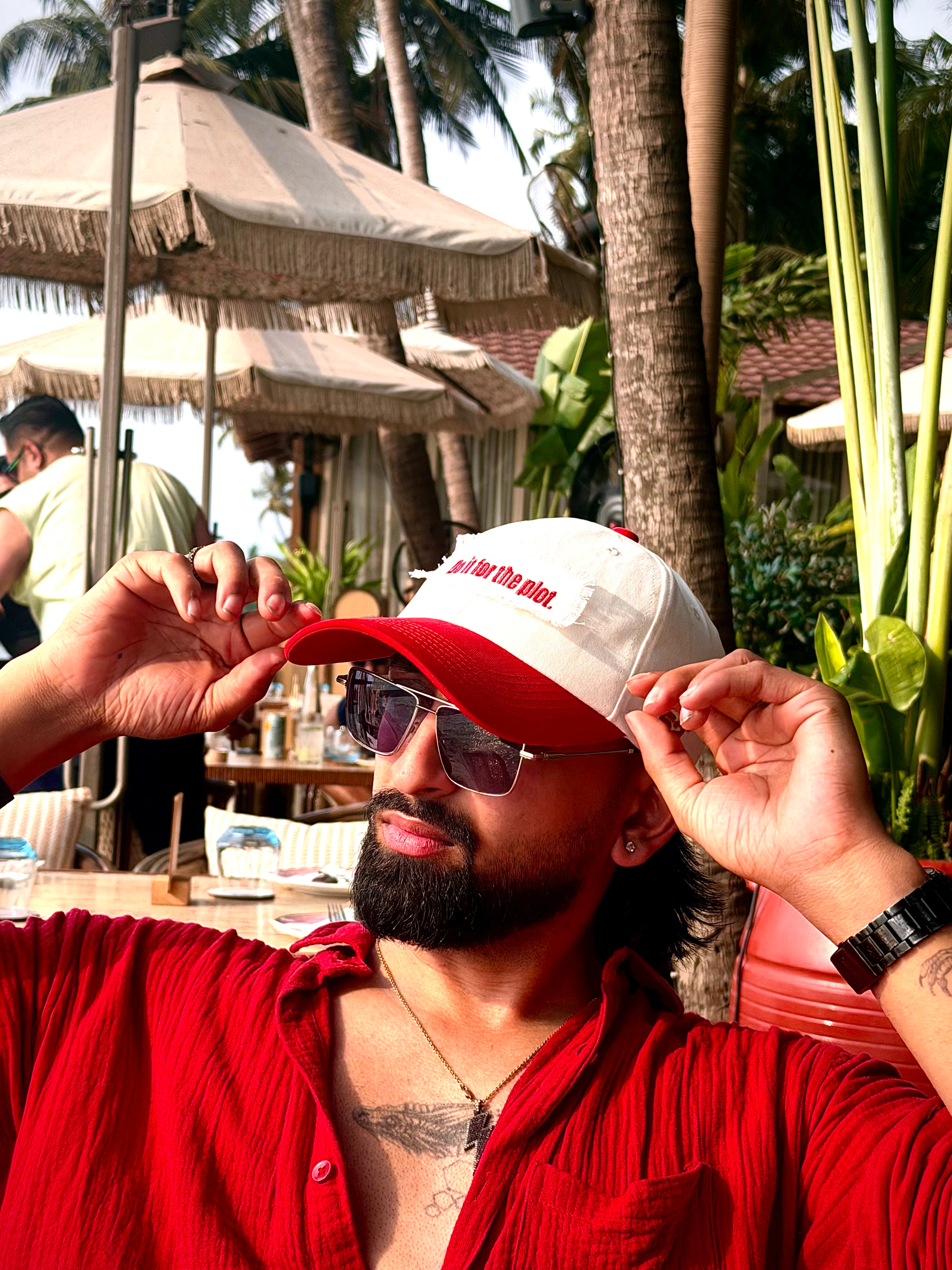 Man wearing a red and white cap with sunglasses, sitting outdoors with palm trees and umbrellas in the background.