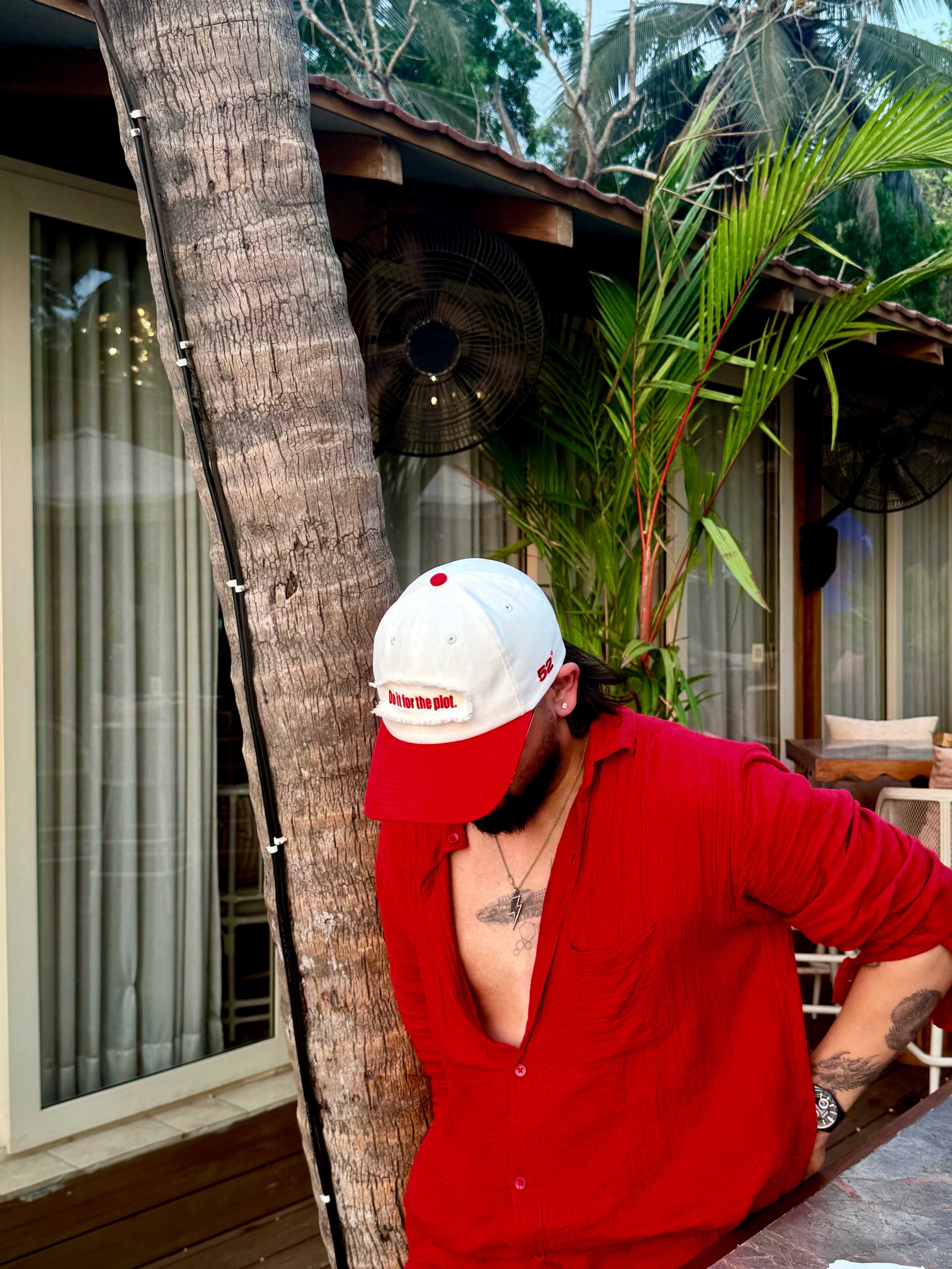 Person wearing a red shirt and white cap leaning against a palm tree in a tropical setting.