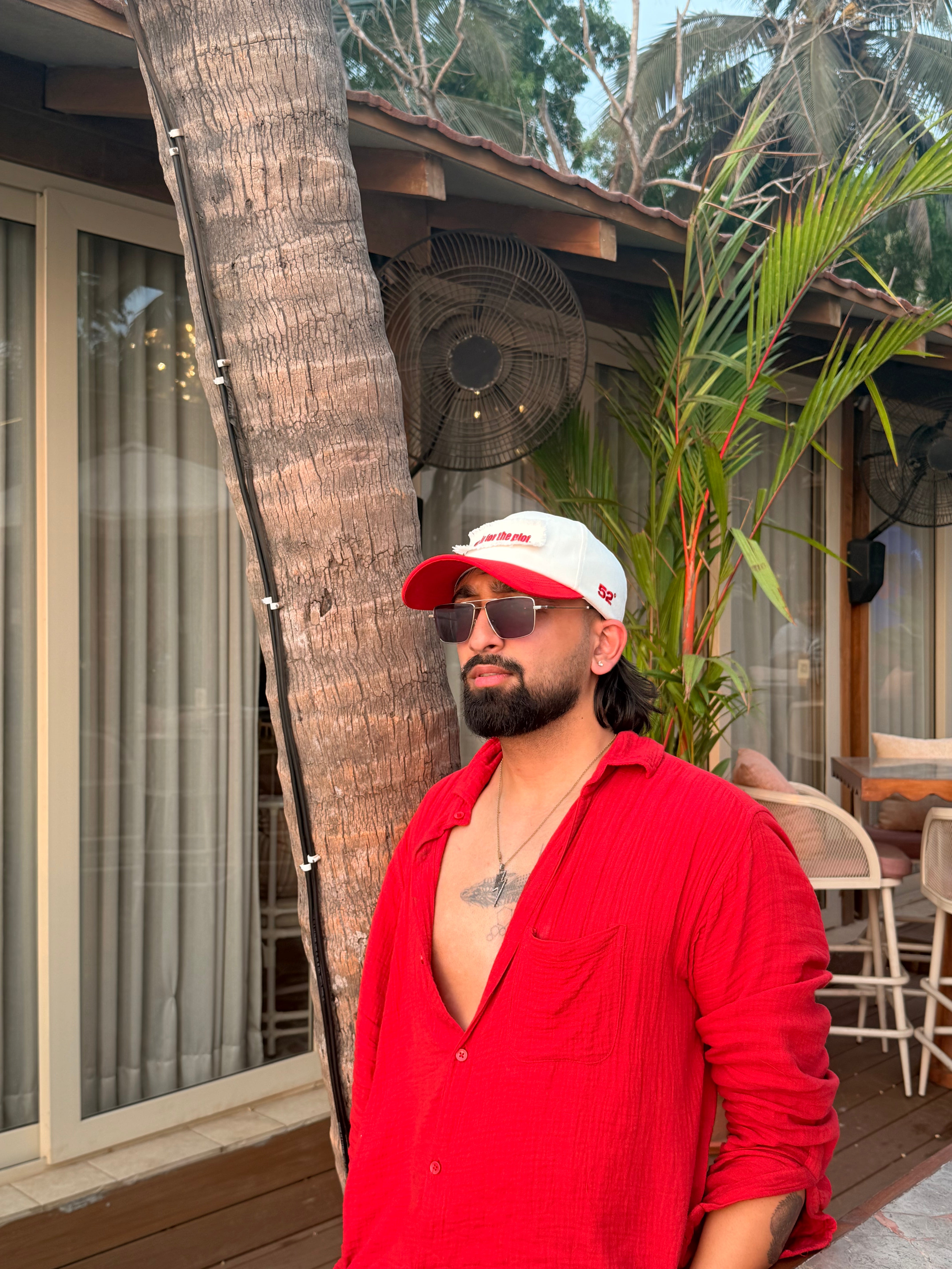 Man wearing a red shirt and white cap standing outdoors near a palm tree