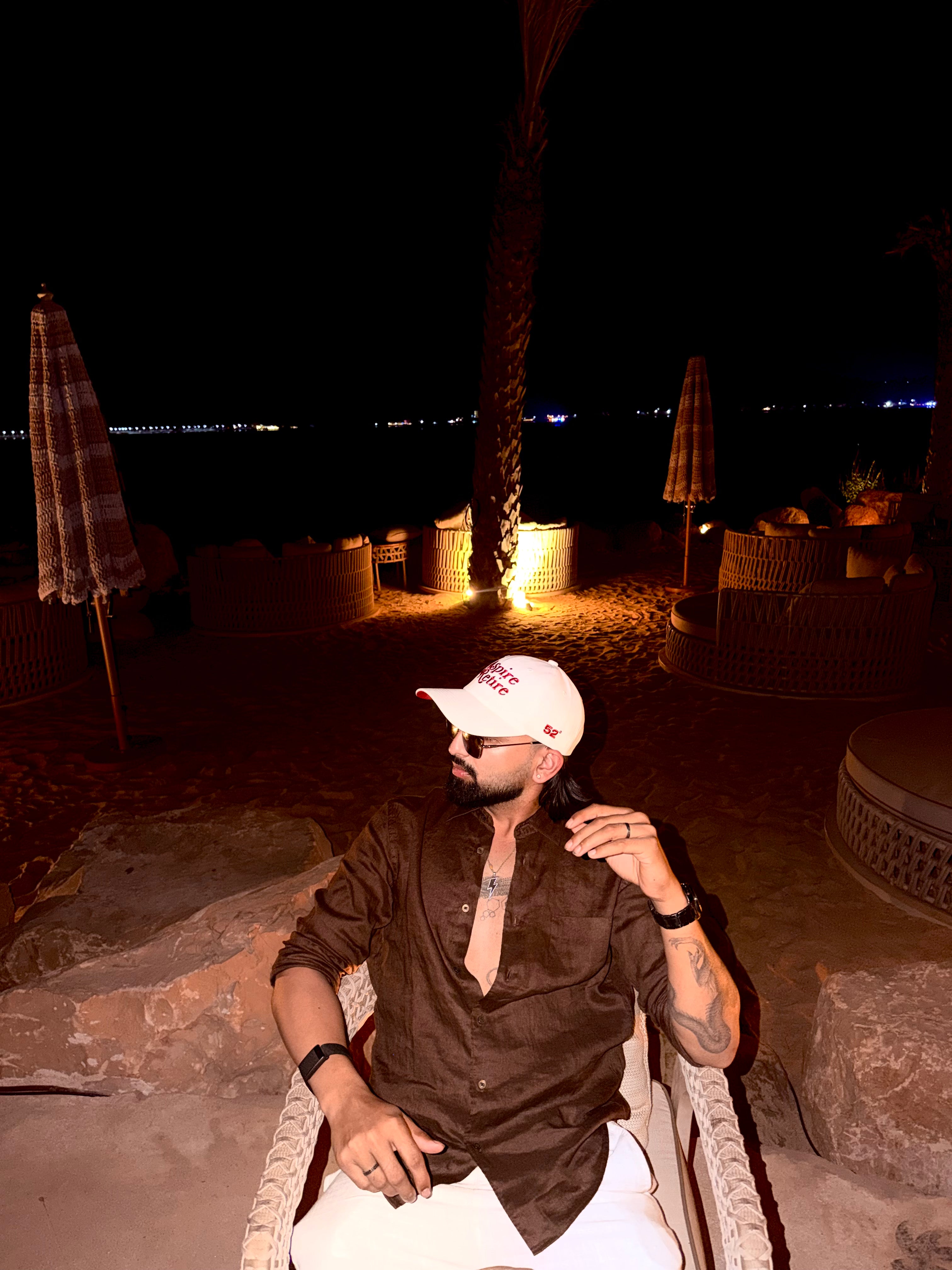 Man sitting on a chair at night with palm trees and lights in the background wearing a cap
