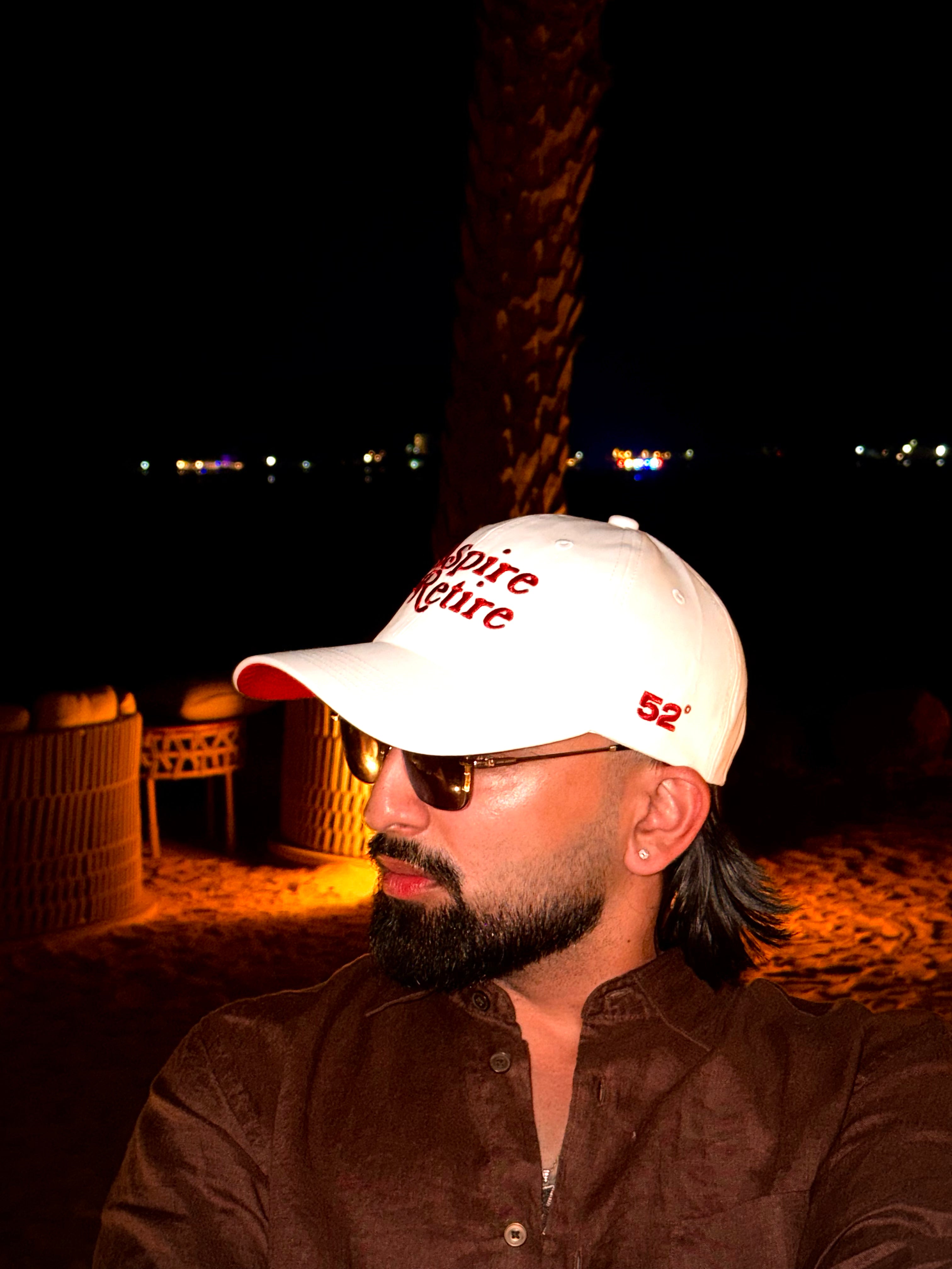 Man wearing a white cap with red text and sunglasses, sitting outdoors at night.