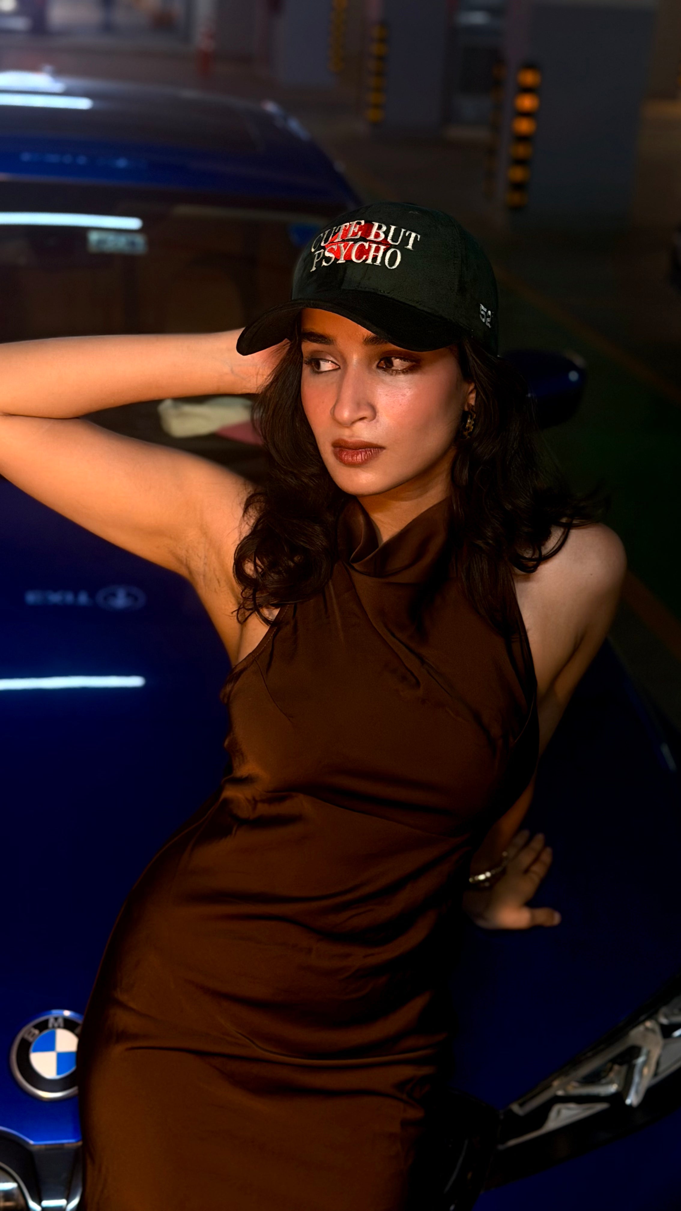Woman in a brown dress and green cap leaning against a blue car at night.