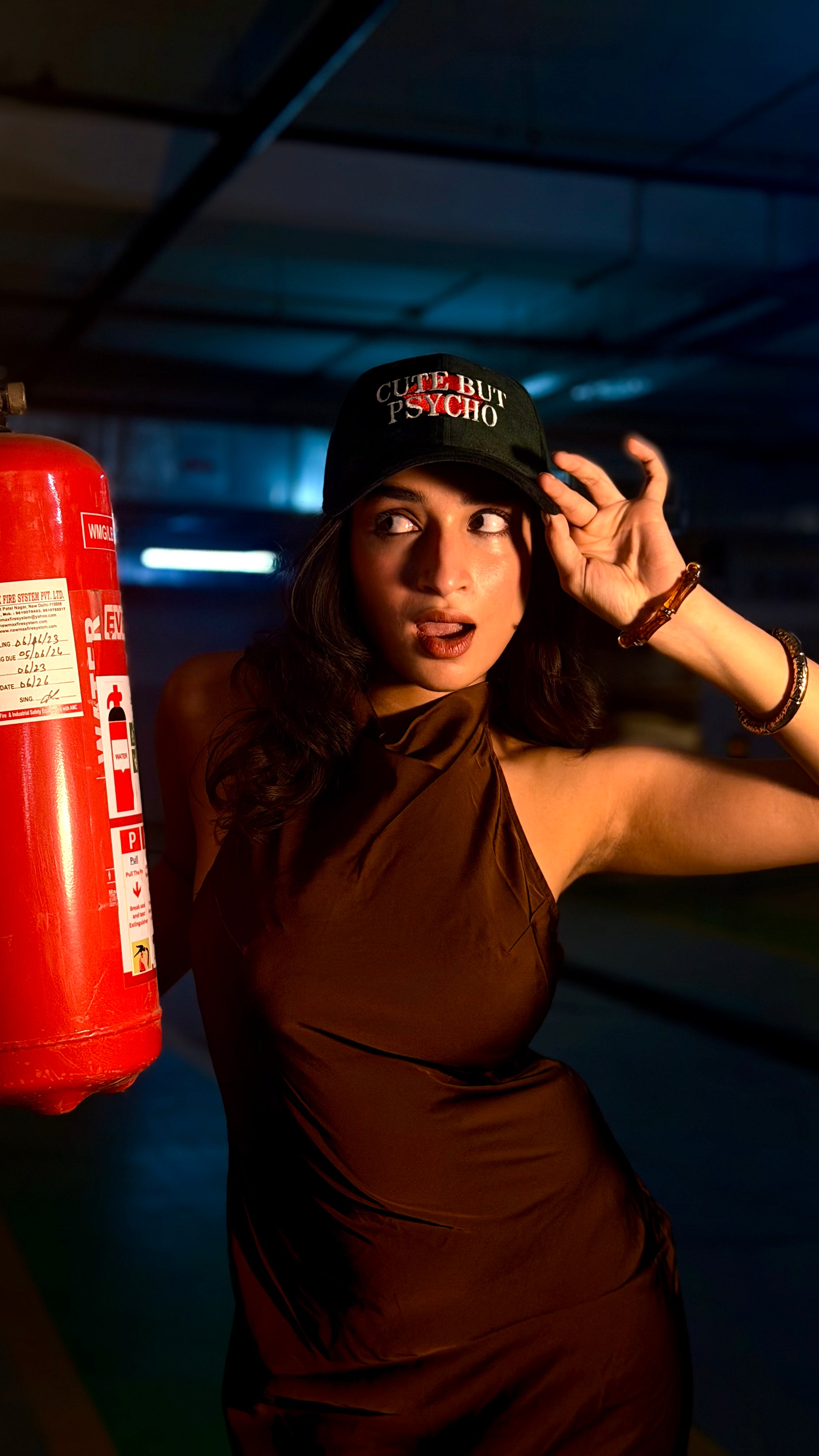 Person wearing a cap and brown dress standing next to a red fire extinguisher in a dimly lit room.