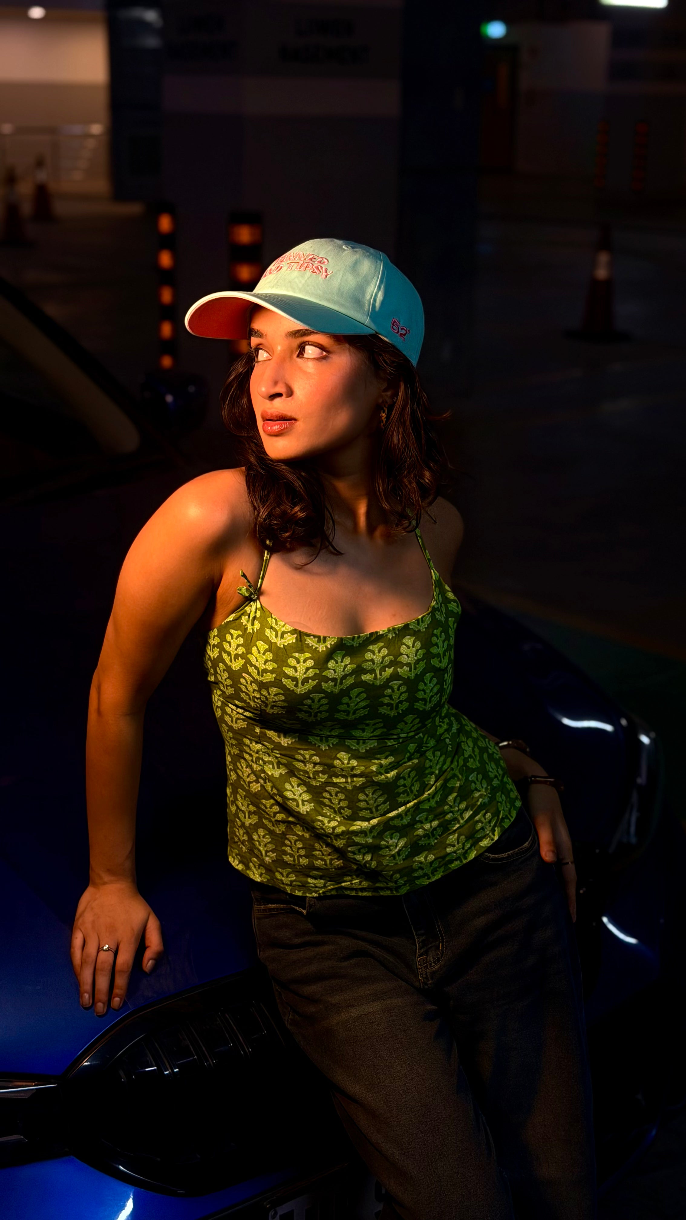 Woman wearing a green patterned top and mint cap sitting on a vehicle at night.