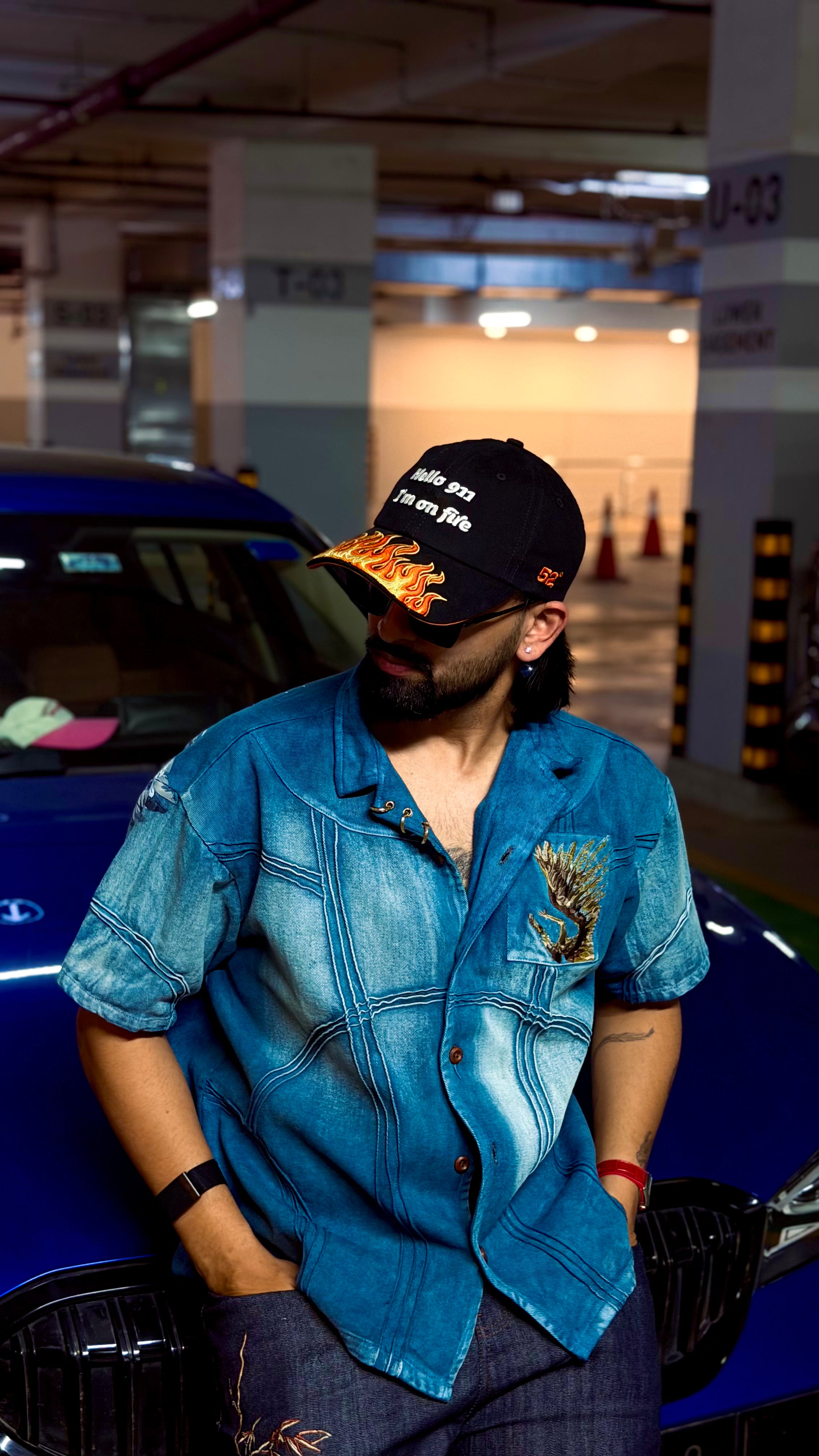 Person wearing a blue denim shirt and black cap standing next to a blue car in an indoor setting.