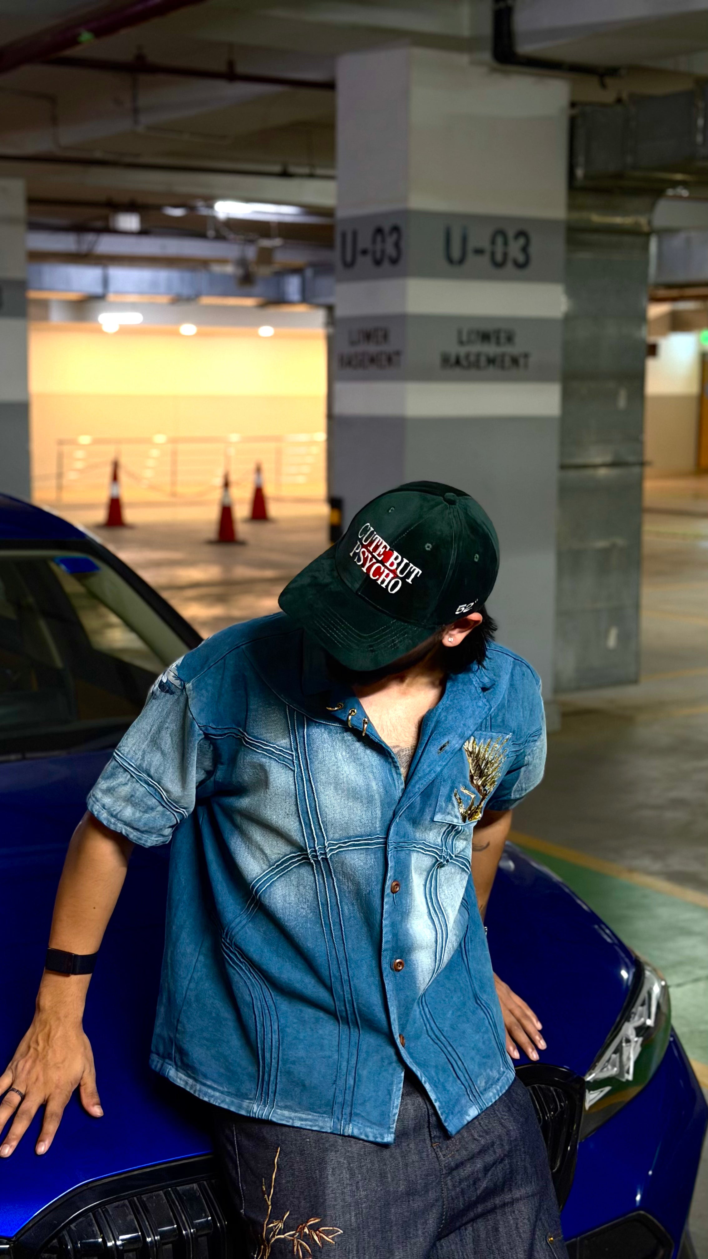 Person wearing a denim jacket and cap leaning against a blue car in a parking garage.