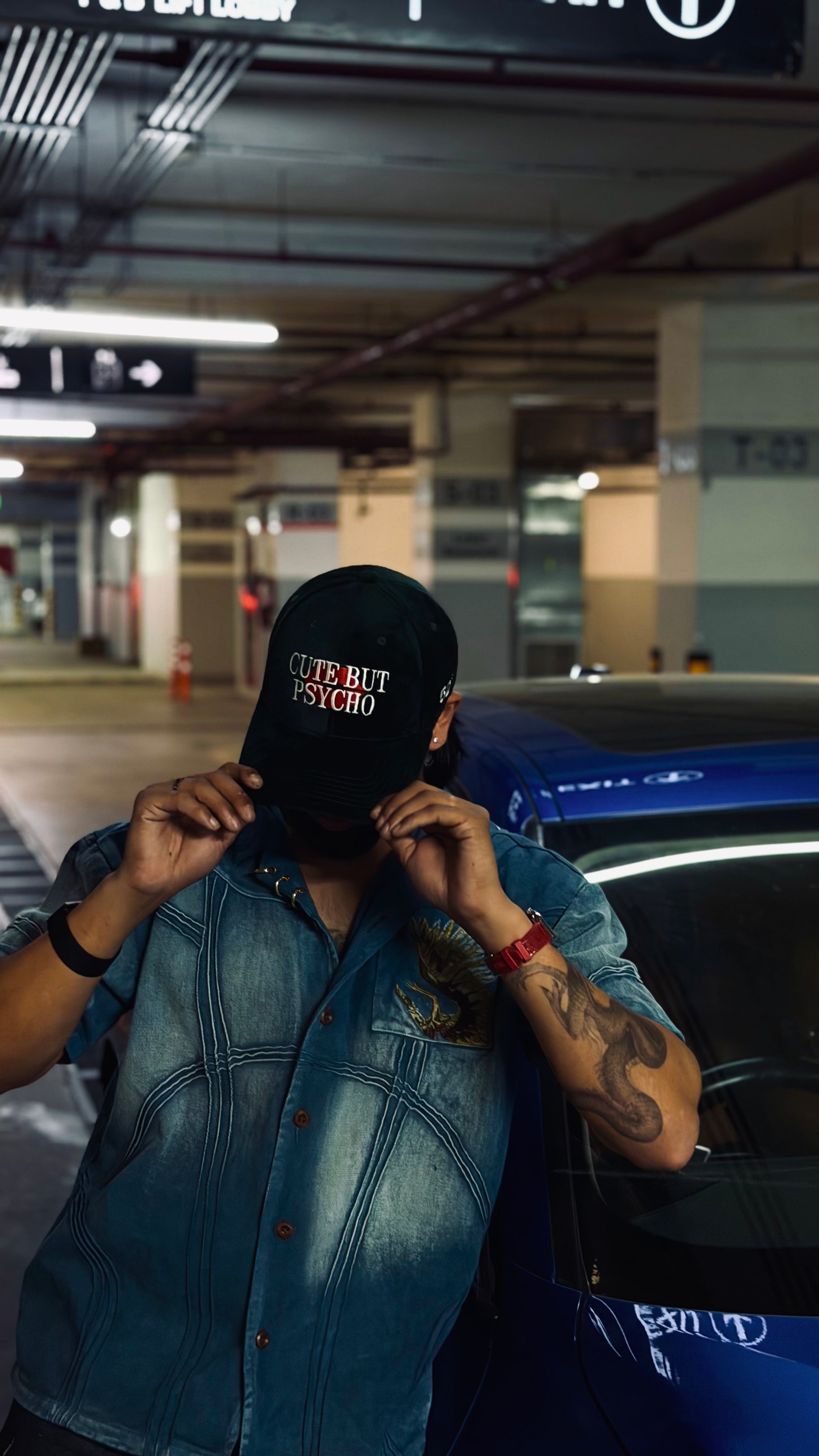Person wearing a denim jacket and green cap with text, standing in a parking garage.