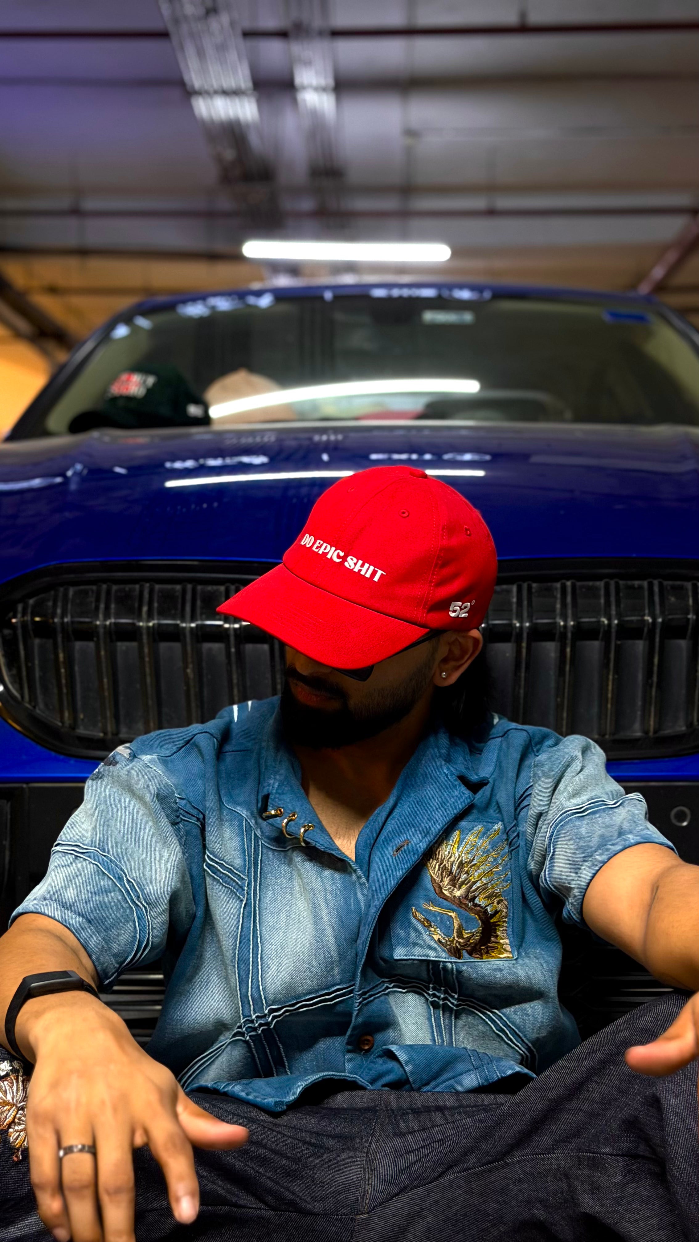Person wearing a red cap and denim jacket sitting in front of a blue car.