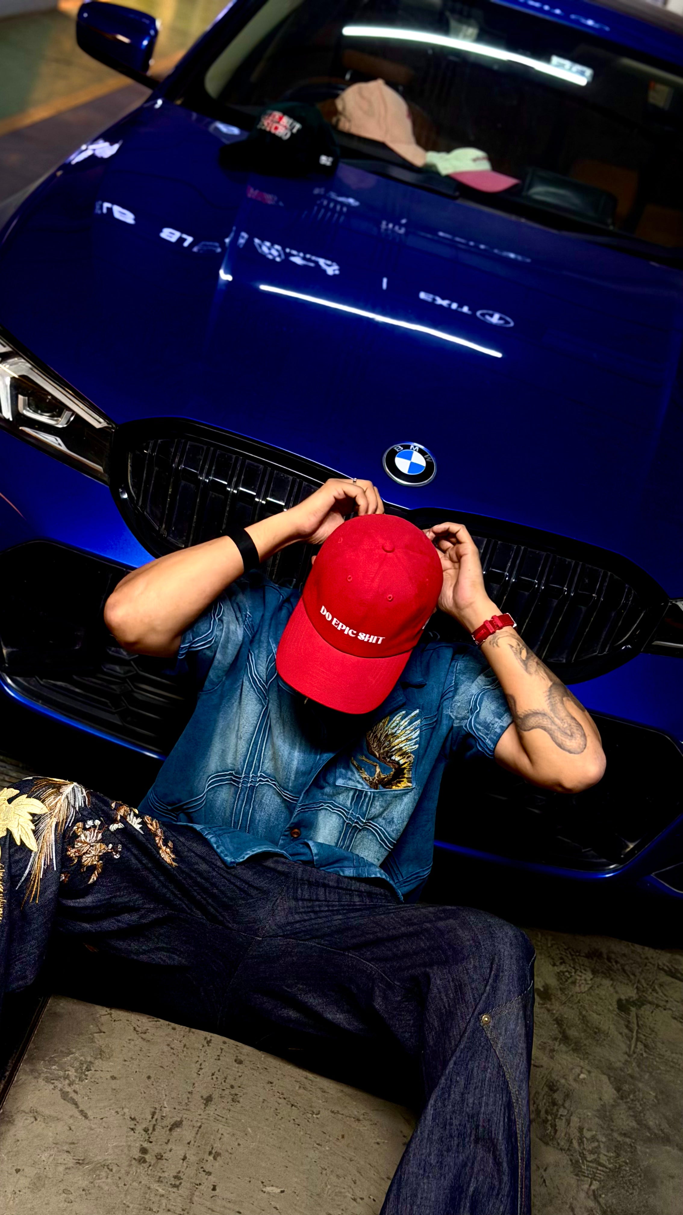 Person wearing a red cap and blue shirt sitting next to a blue BMW car.