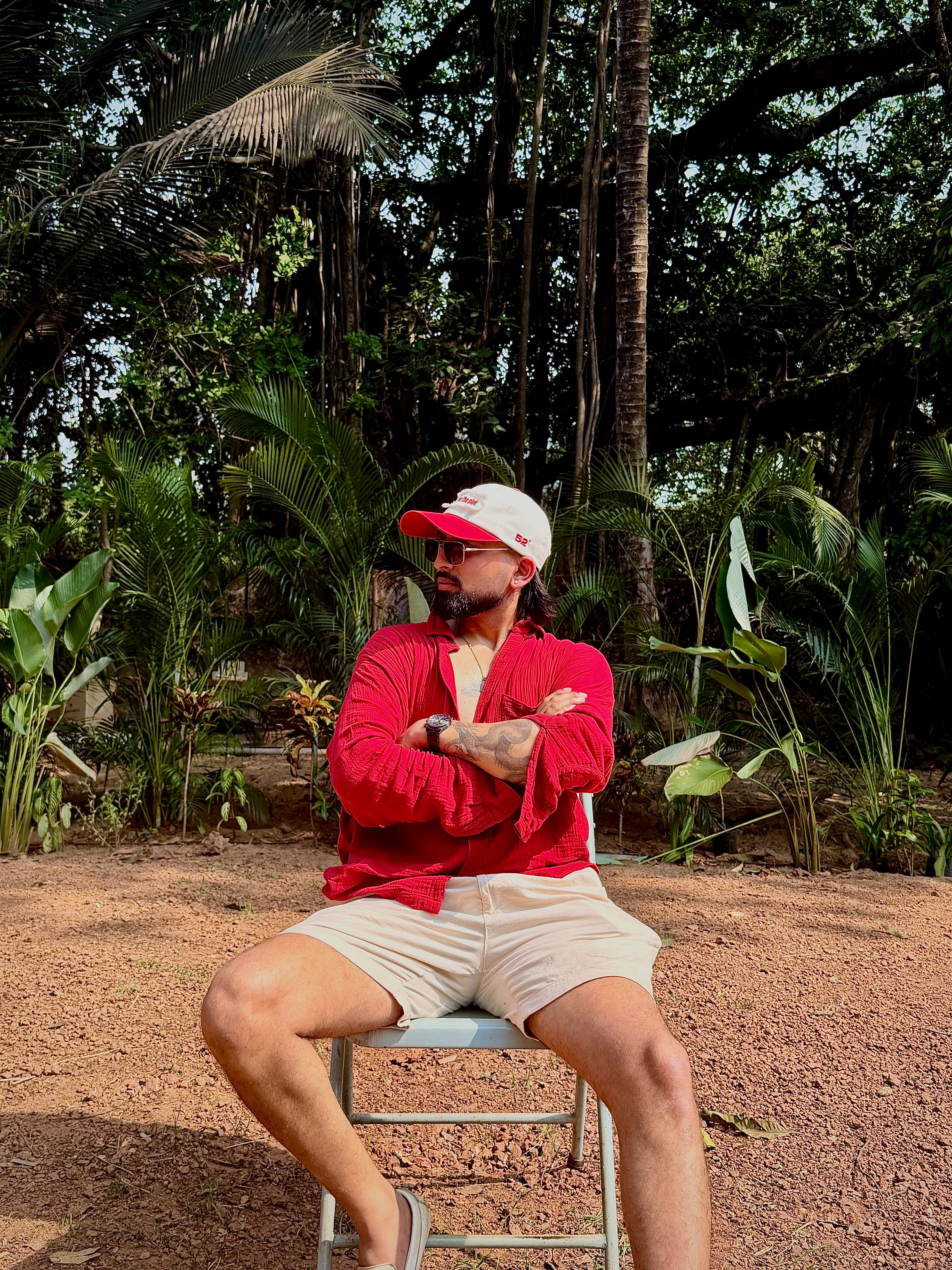 Man in red shirt and white cap sitting on a chair in a tropical setting with palm trees wearing do it for the plot cap