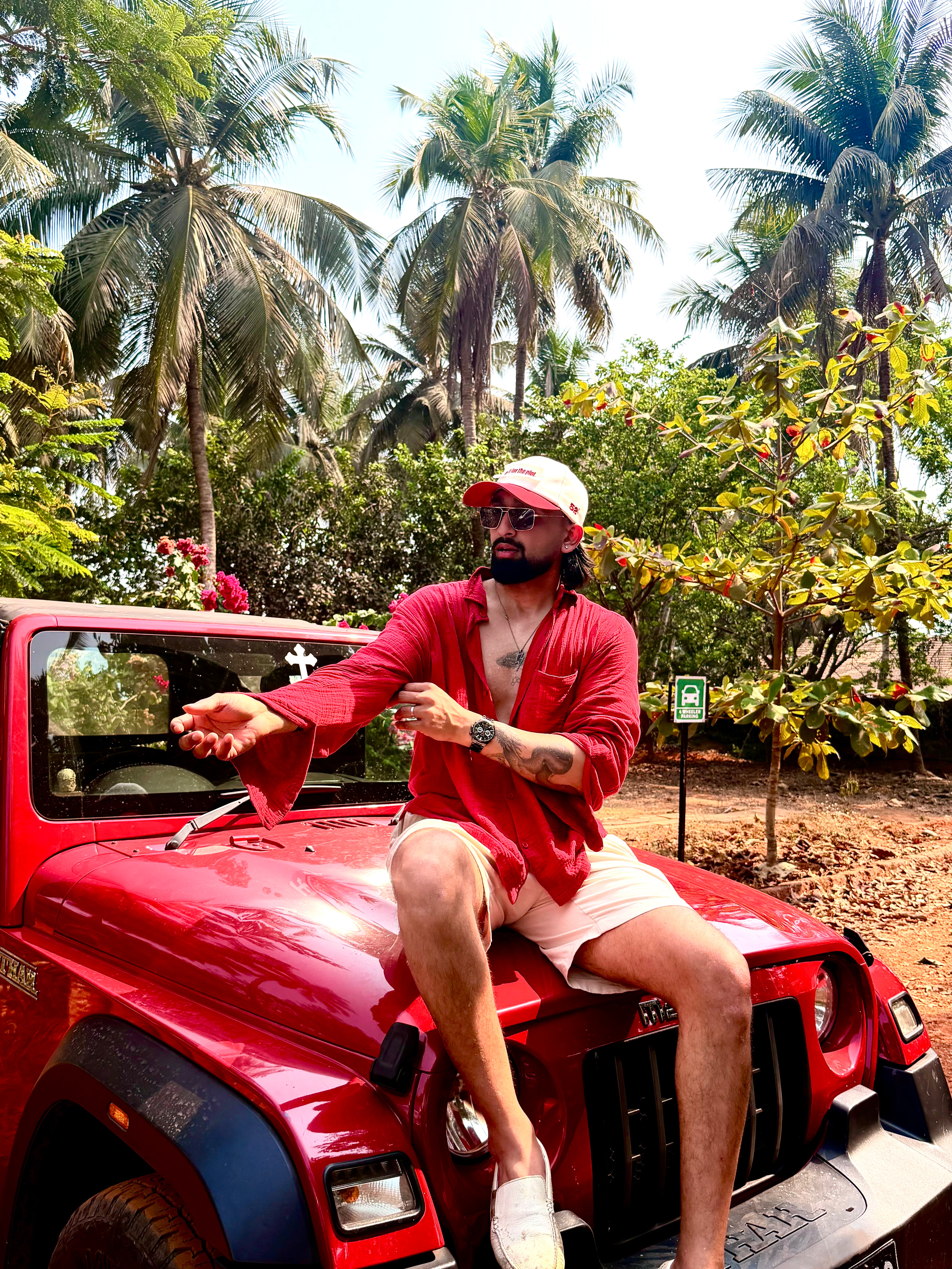 Man sitting on a red jeep with palm trees and greenery in the background wearing do it for the plot cap