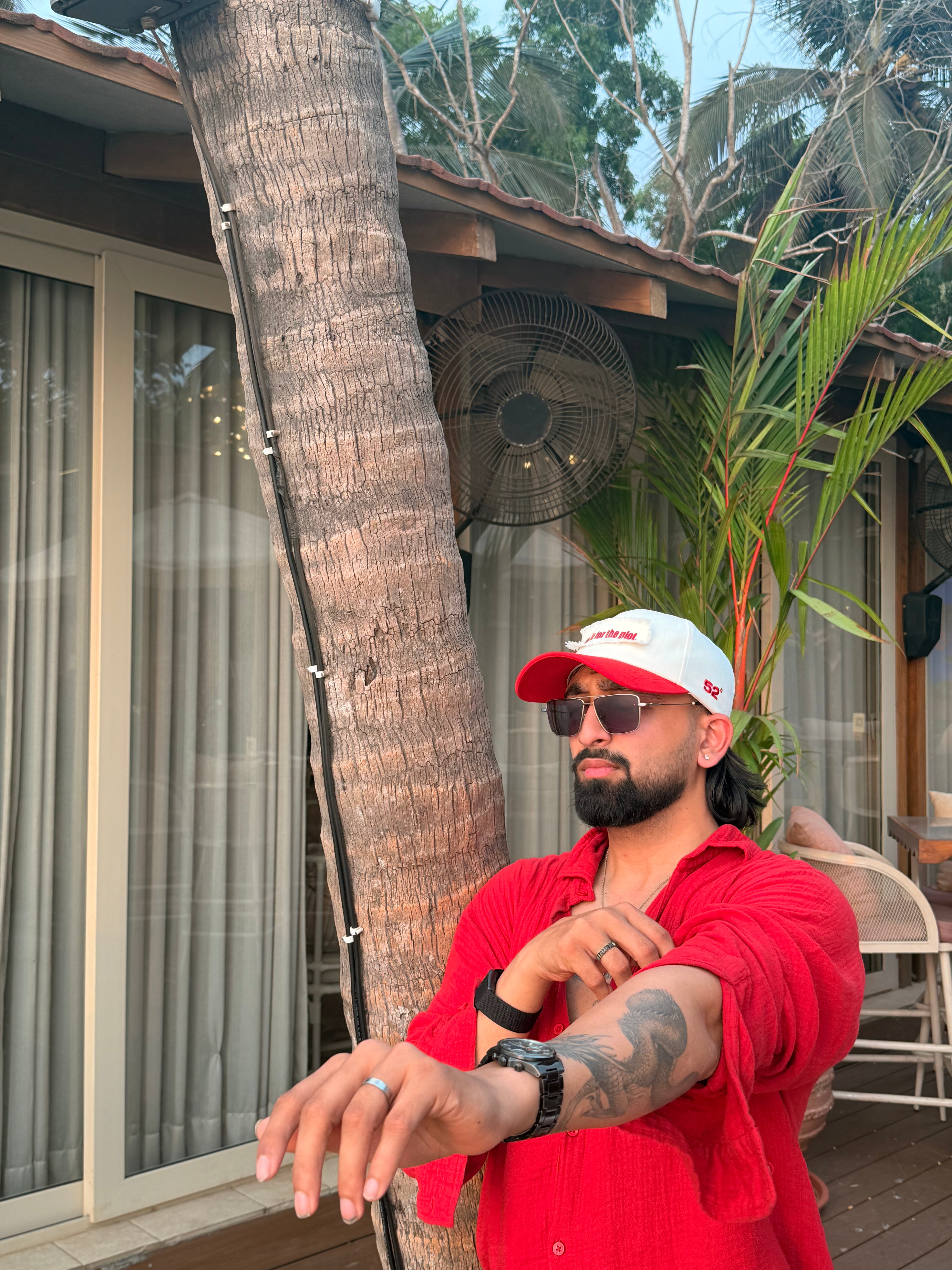 Man in a red shirt and white cap sitting outdoors with palm trees and a building in the background