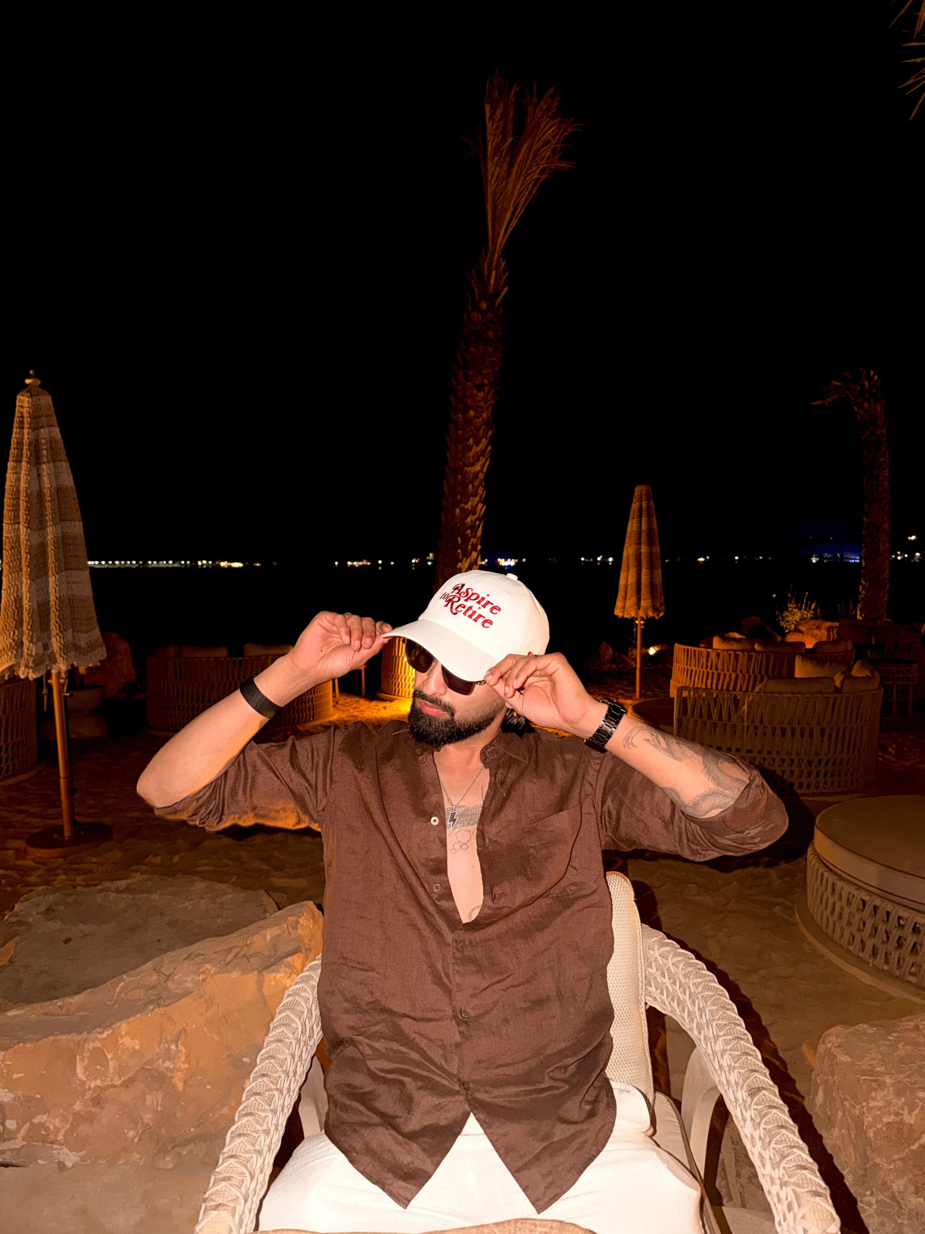 Man adjusting a cap at an outdoor nighttime setting with palm trees and umbrellas.