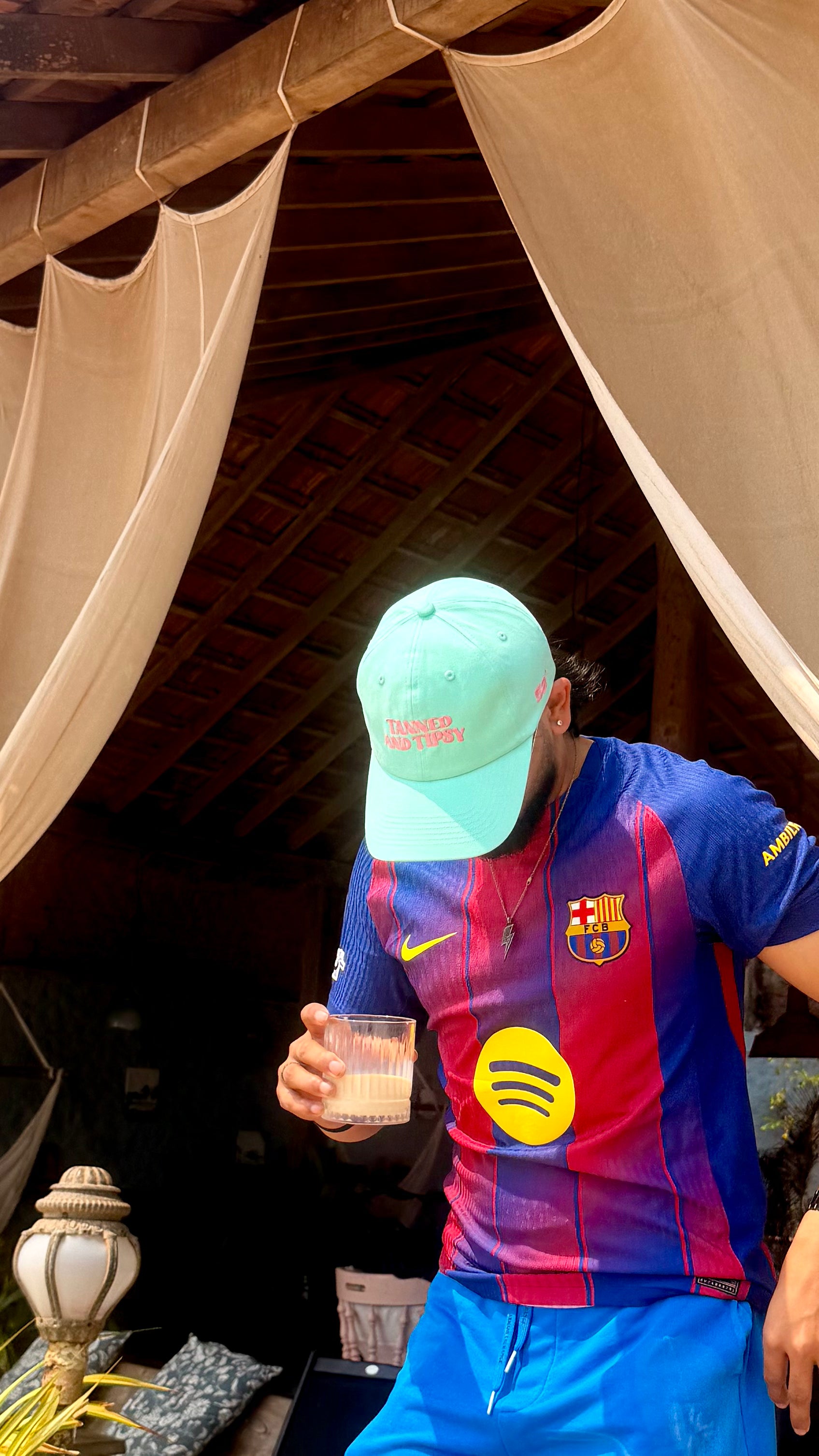 Person wearing a colorful sports jersey and light blue cap, holding a glass, under a canopy.