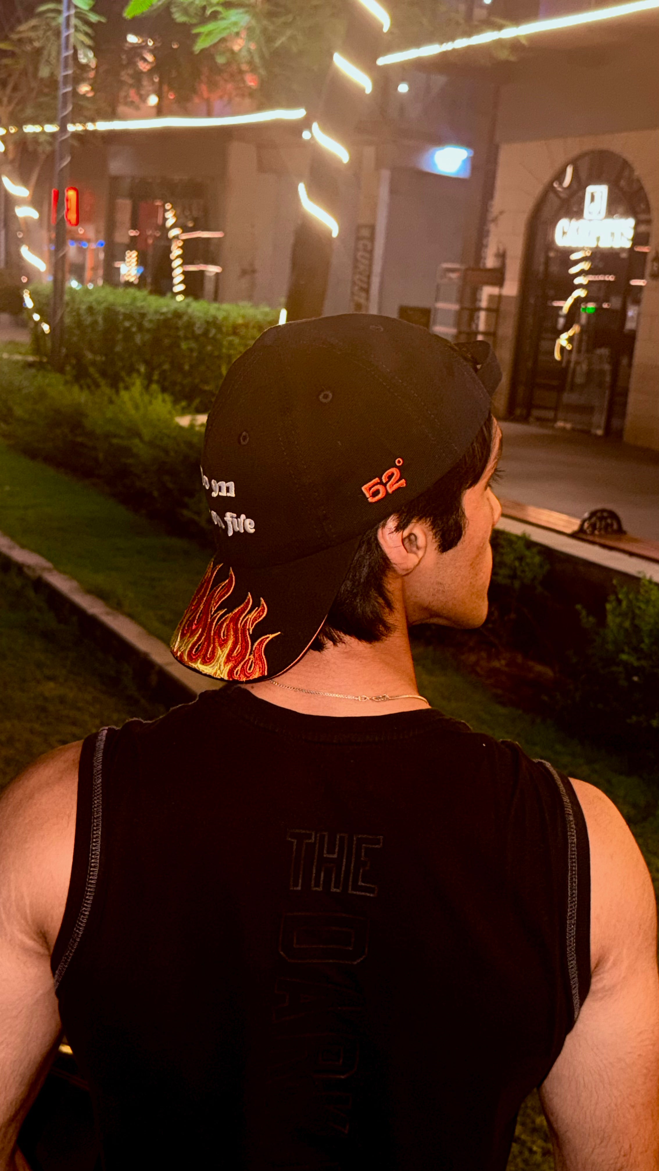 Person wearing a black cap with flame design and 'THC' tank top in an urban setting at night.