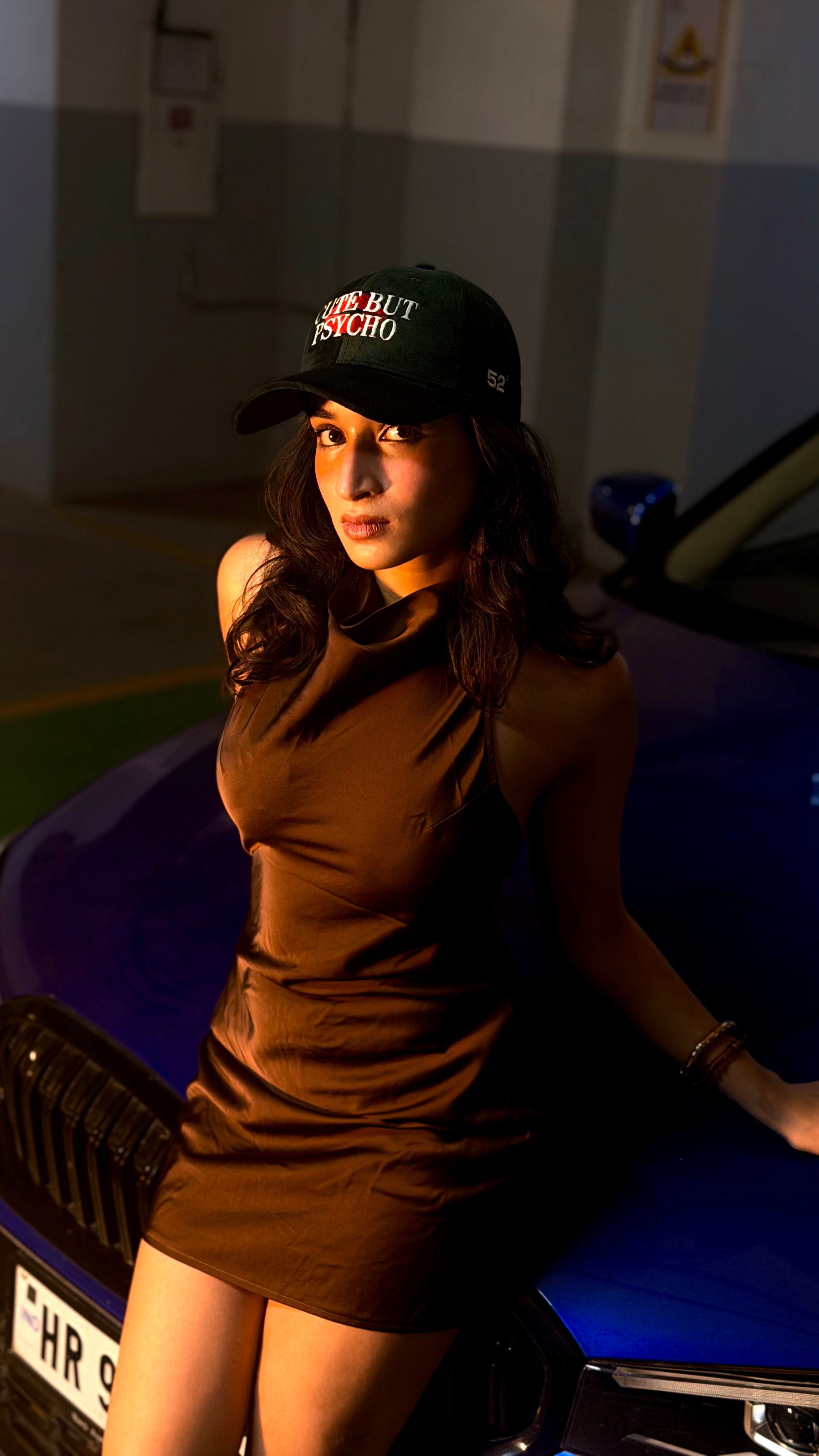 Woman in a brown dress and green  cap leaning against a blue car in a garage.