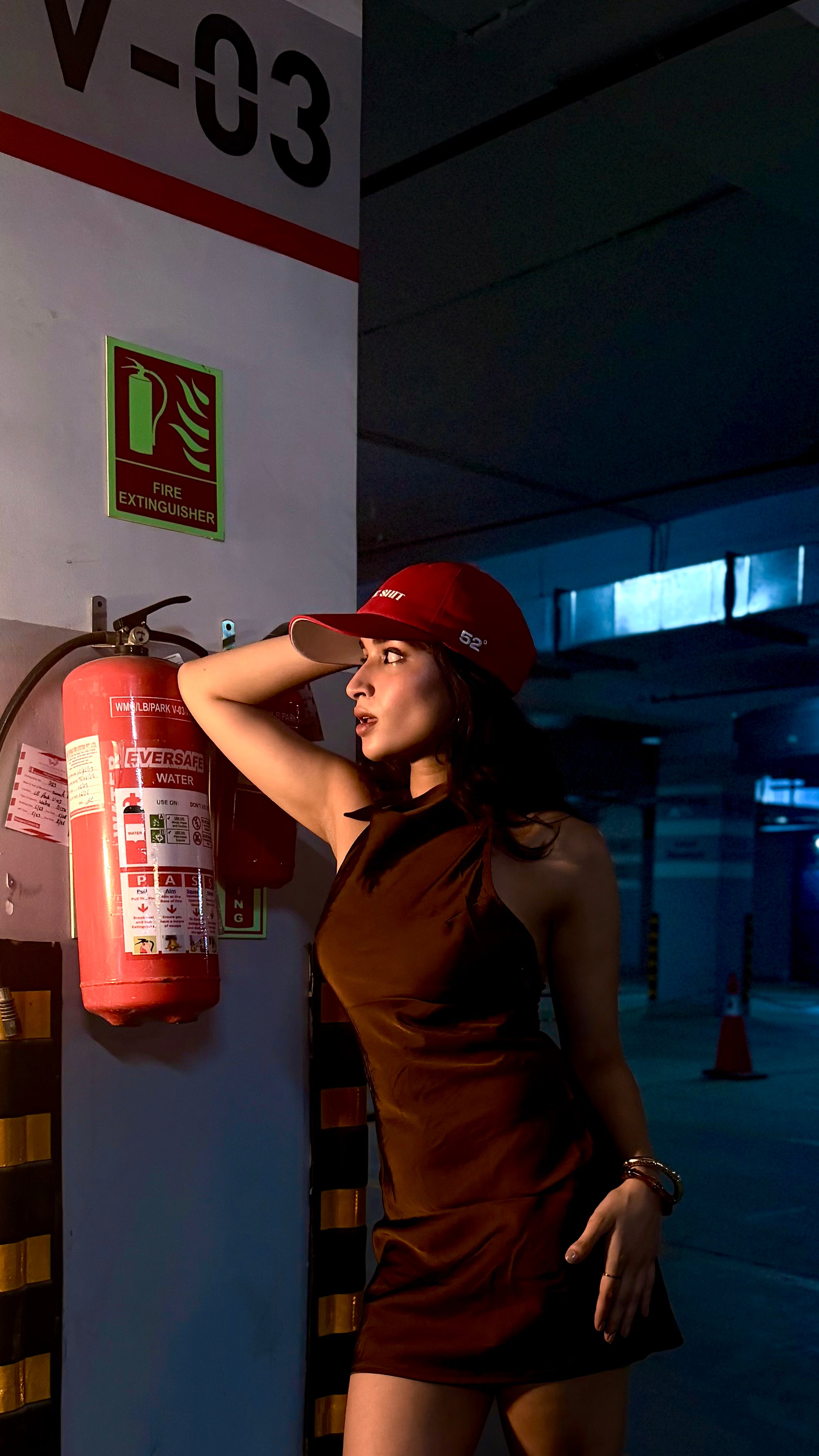 Woman in a red cap and dress standing next to a fire extinguisher in a dimly lit area.
