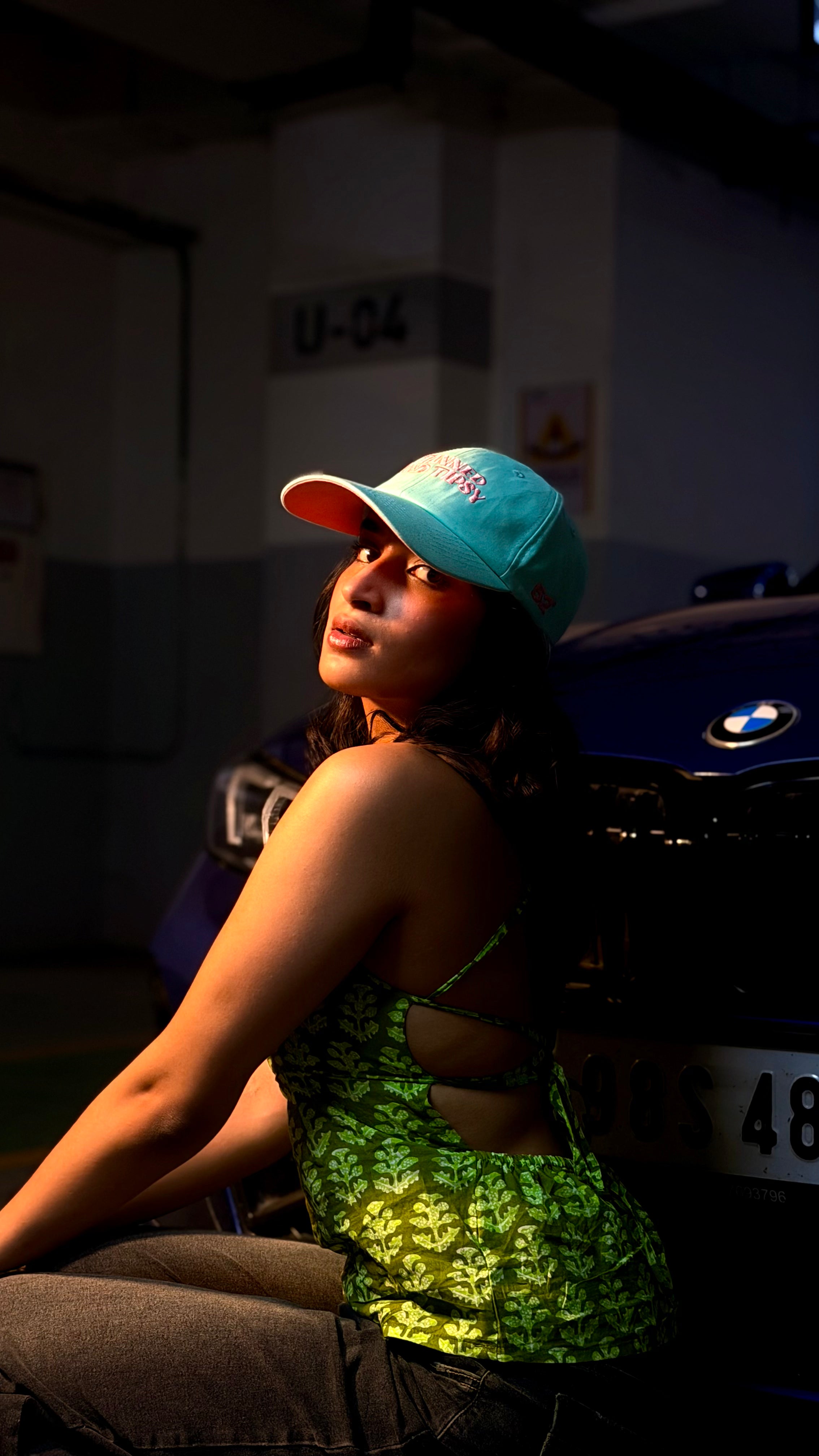 Person wearing a blue cap and green top sitting next to a car in a dark setting